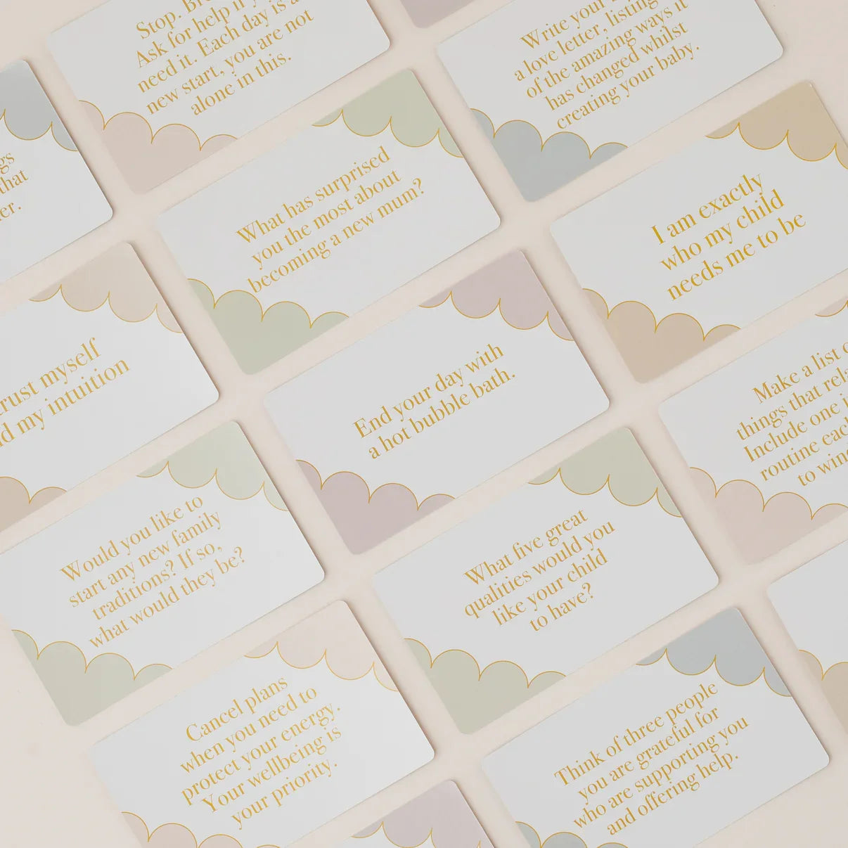A close-up of multiple cream-colored cards with scalloped edges, printed with soft pastel text and gold lettering, arranged diagonally on a light beige background.