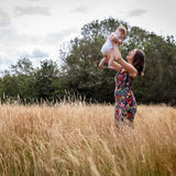 A woman in a colorful floral dress stands in a field of tall dry grass, lifting a baby into the air outdoors with trees and a cloudy sky in the background.