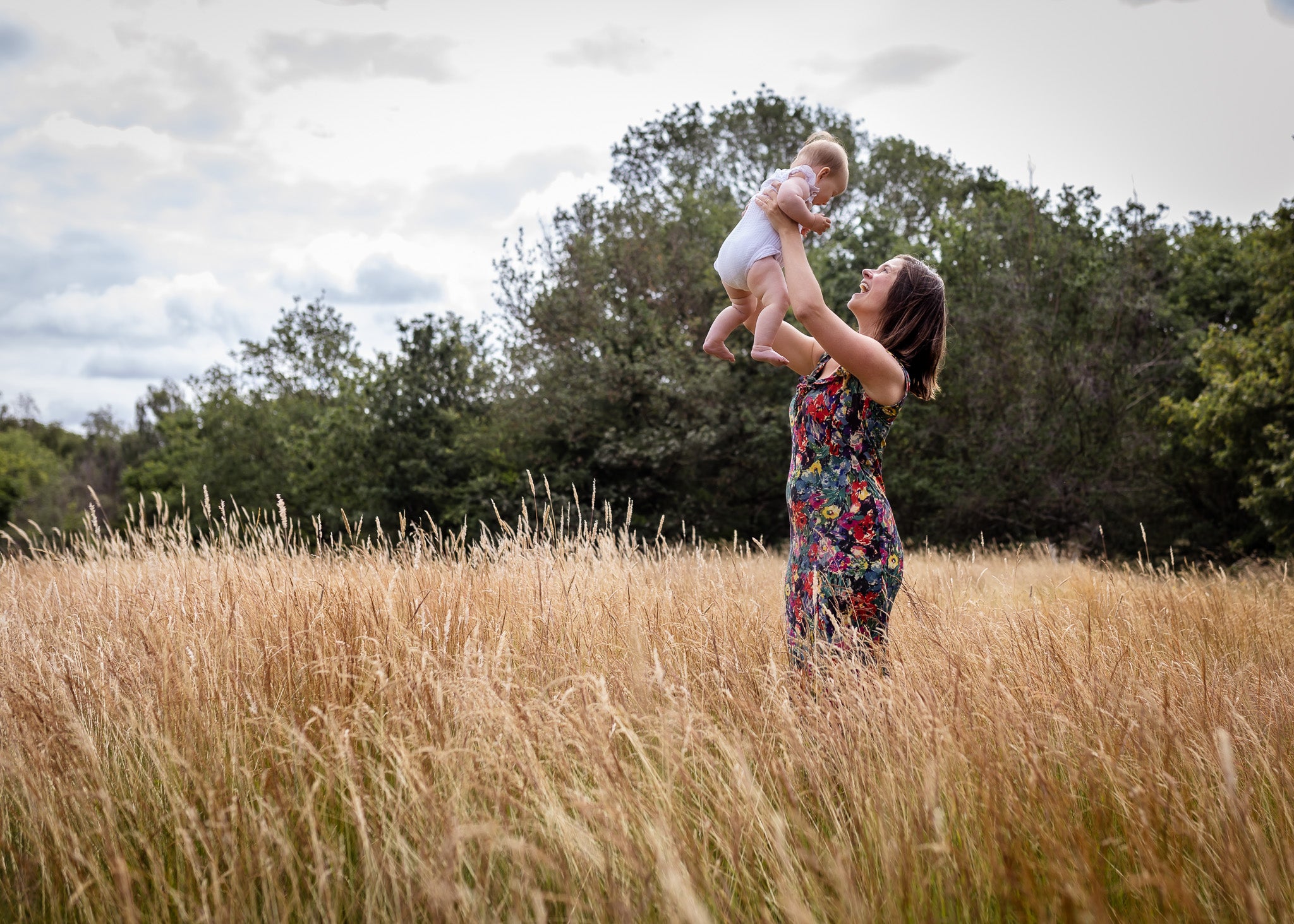 A woman in a colorful floral dress stands in a field of tall dry grass, lifting a baby into the air outdoors with trees and a cloudy sky in the background.