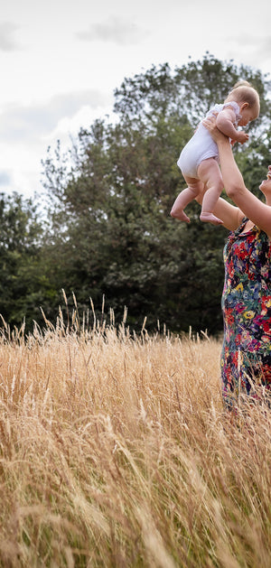 A woman in a colorful floral dress stands in a field of tall dry grass, lifting a baby into the air outdoors with trees and a cloudy sky in the background.