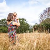 A woman in a colorful floral dress holds a baby in a tall golden field under a blue sky with clouds.
