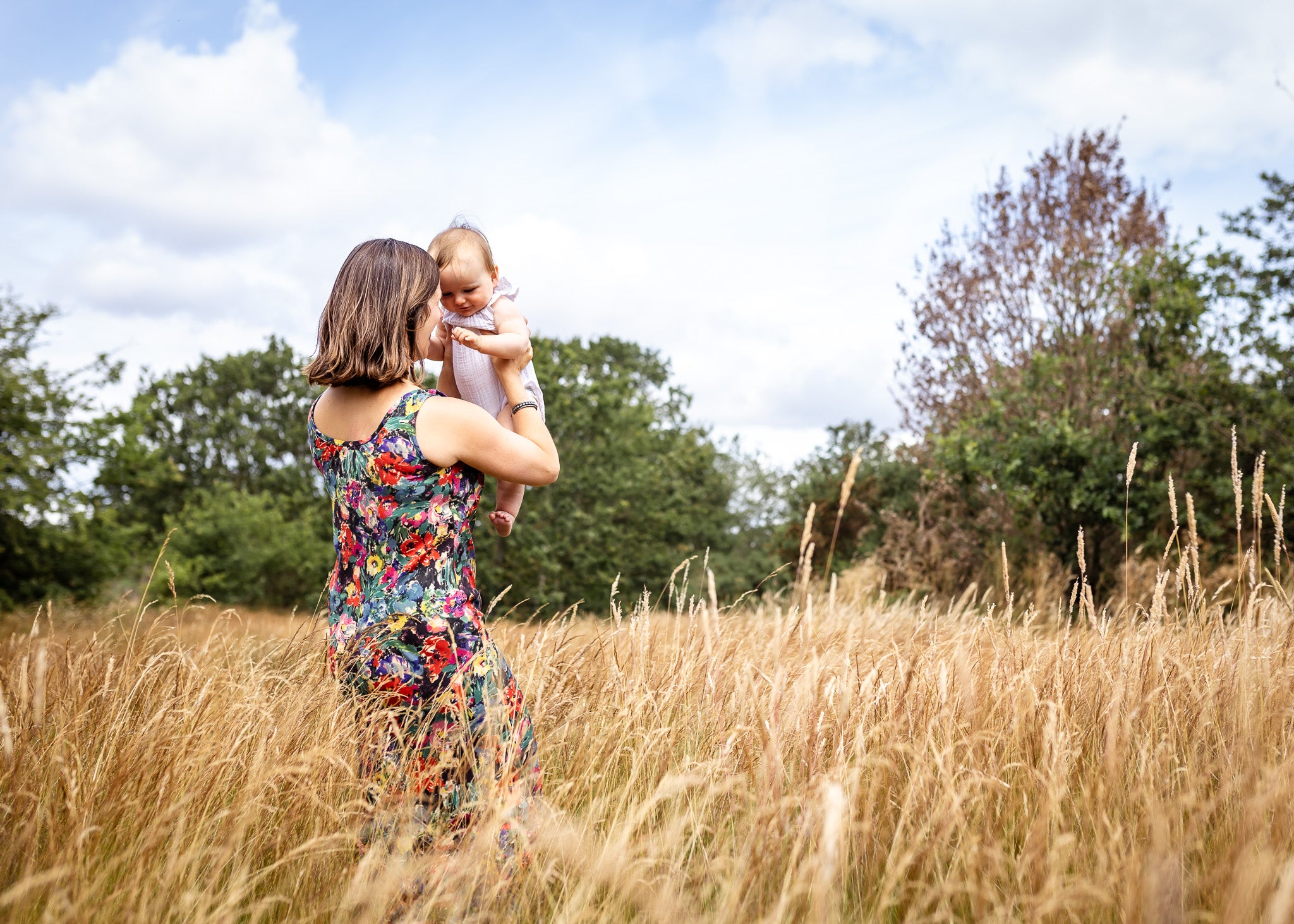 A woman in a colorful floral dress holds a baby in a tall golden field under a blue sky with clouds.