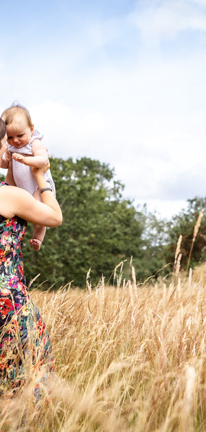 A woman in a colorful floral dress holds a baby in a tall golden field under a blue sky with clouds.