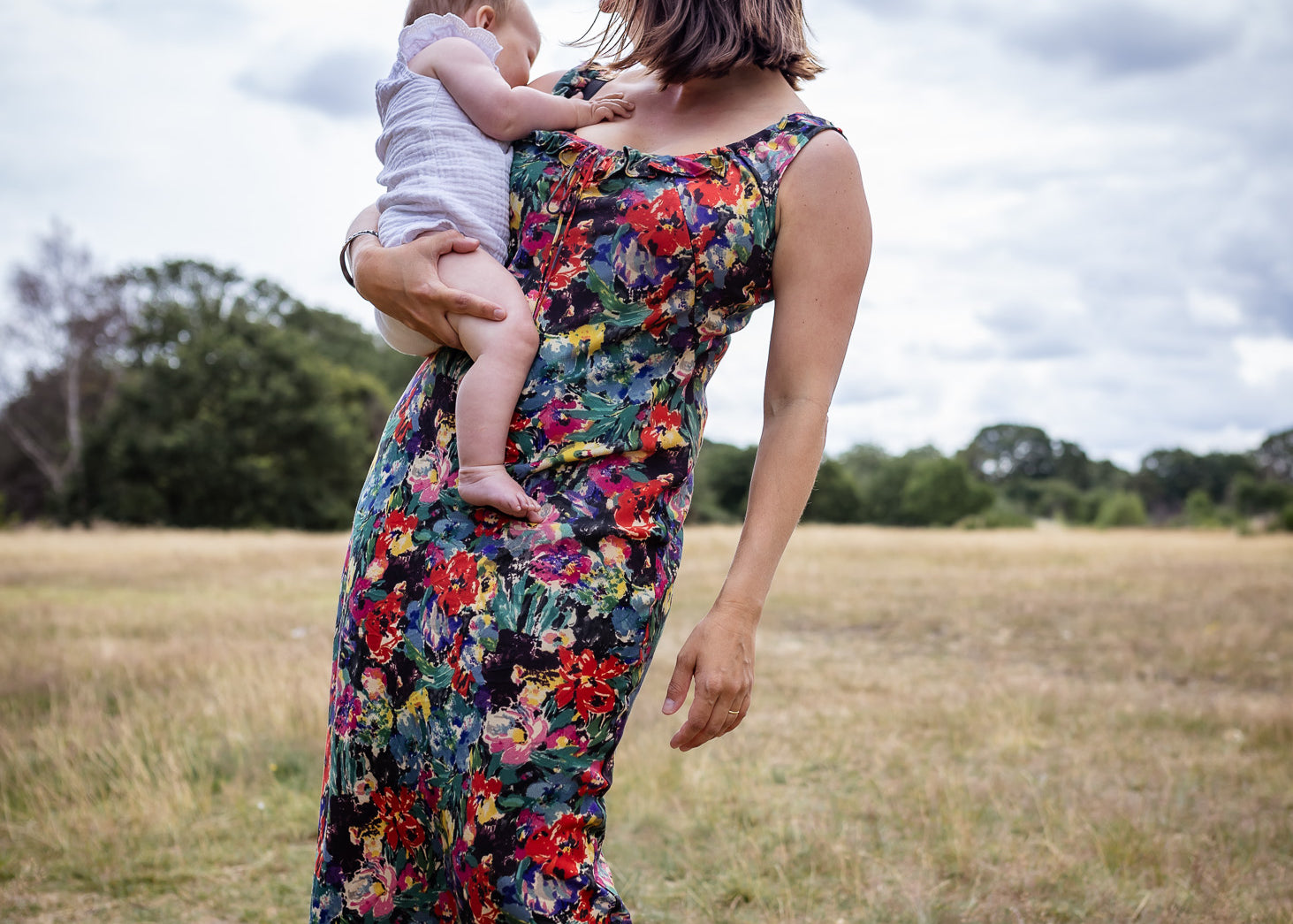A woman standing in an open grassy field under a cloudy sky, holding a baby. She is wearing a sleeveless maxi dress with a dense multicolor floral print on a dark background.