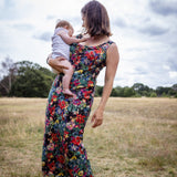 A woman standing in an open grassy field under a cloudy sky, holding a baby. She is wearing a sleeveless maxi dress with a dense multicolor floral print on a dark background.