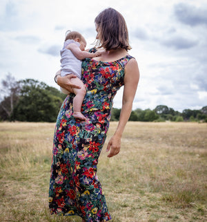 A woman standing in an open grassy field under a cloudy sky, holding a baby. She is wearing a sleeveless maxi dress with a dense multicolor floral print on a dark background.