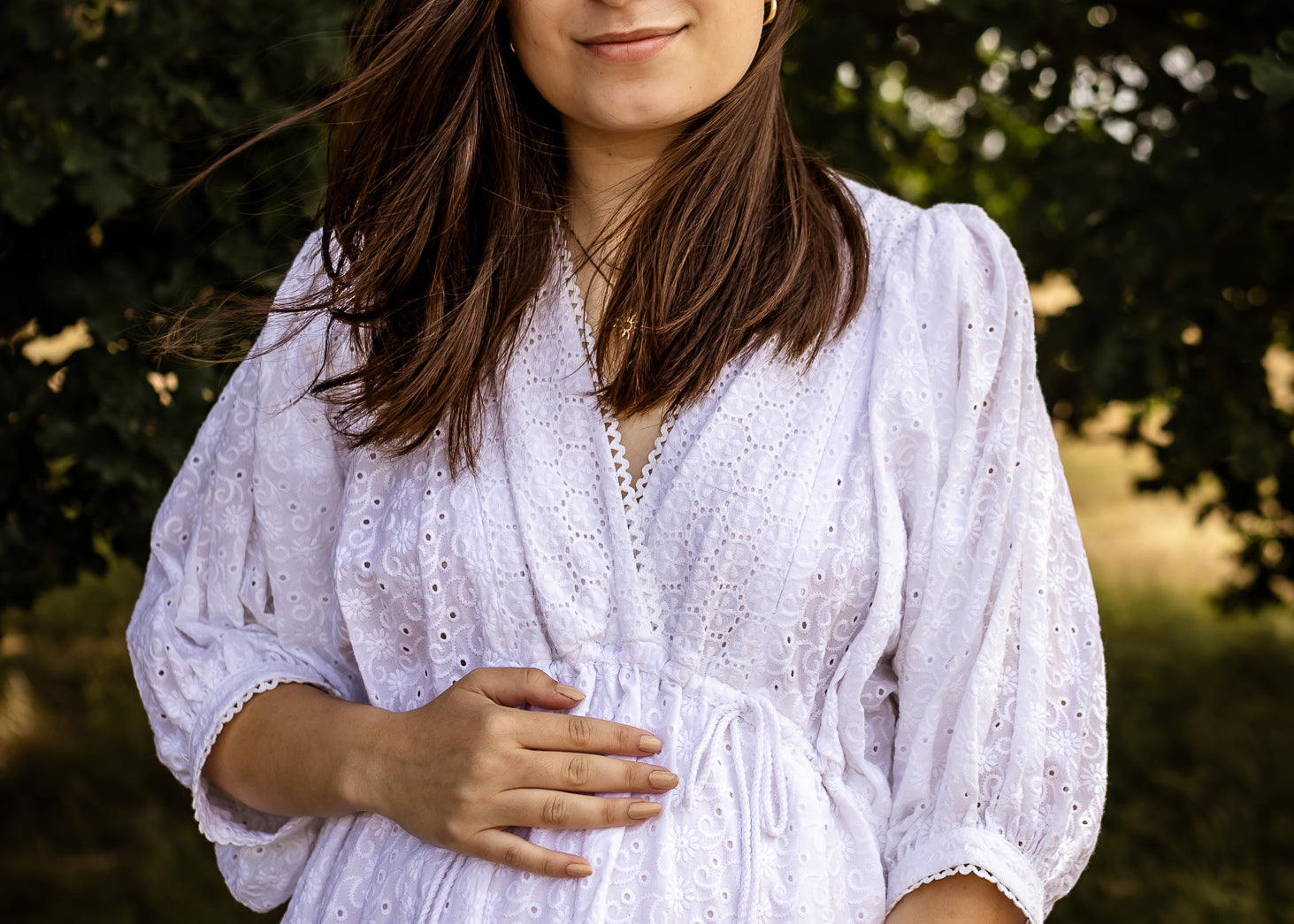 A pregnant woman wearing a white eyelet long-sleeve maternity dress, standing outdoors in front of a soft-focus background of green trees.
