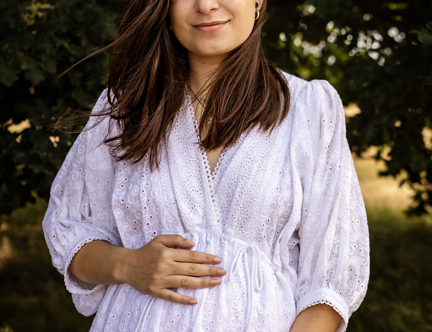 A pregnant woman wearing a white eyelet long-sleeve maternity dress, standing outdoors in front of a soft-focus background of green trees.