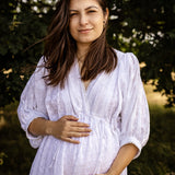 A pregnant woman wearing a white eyelet long-sleeve maternity dress, standing outdoors in front of a soft-focus background of green trees.