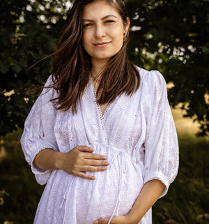 A pregnant woman wearing a white eyelet long-sleeve maternity dress, standing outdoors in front of a soft-focus background of green trees.