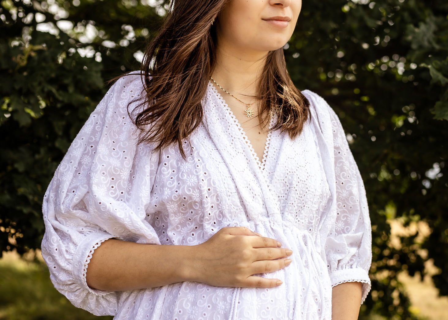 A pregnant woman wearing a white eyelet long-sleeve maternity dress stands outdoors in front of a blurred green tree background, cradling her baby bump.