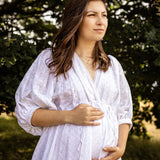A pregnant woman wearing a white eyelet long-sleeve maternity dress stands outdoors in front of a blurred green tree background, cradling her baby bump.