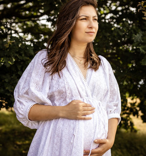 A pregnant woman wearing a white eyelet long-sleeve maternity dress stands outdoors in front of a blurred green tree background, cradling her baby bump.