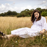A woman sitting in a grassy field wearing a white long-sleeve dress and dark shoes, looking down at the ground with a serene expression.