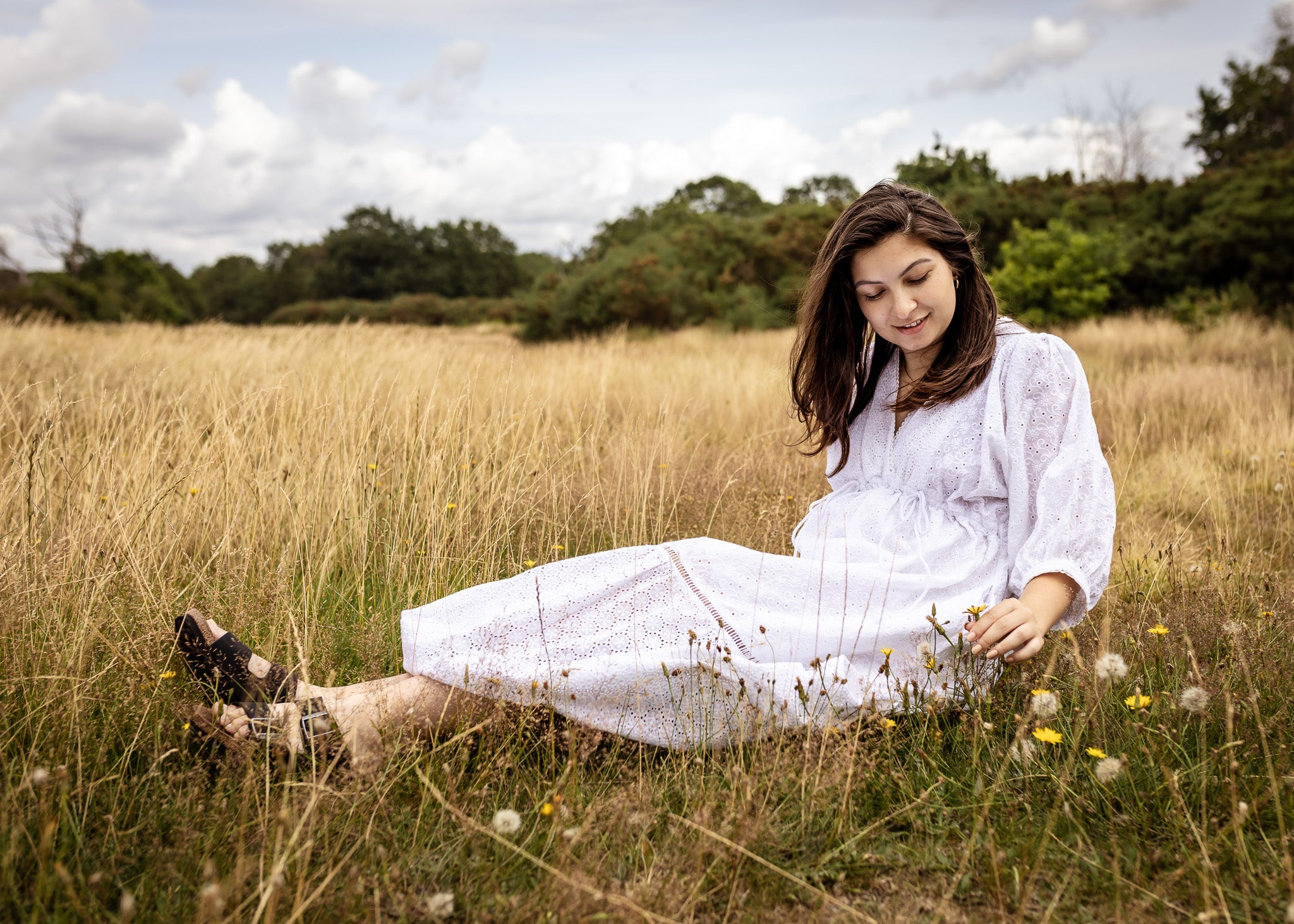 A woman sitting in a grassy field wearing a white long-sleeve dress and dark shoes, looking down at the ground with a serene expression.