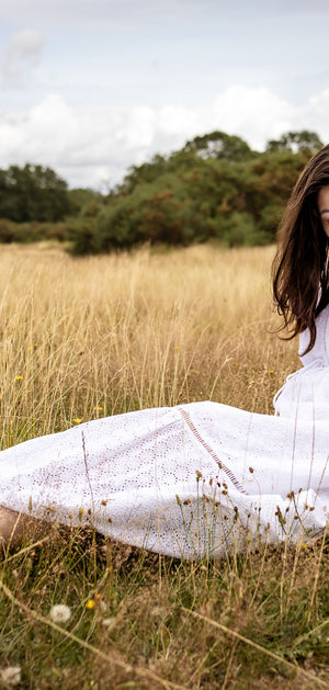 A woman sitting in a grassy field wearing a white long-sleeve dress and dark shoes, looking down at the ground with a serene expression.