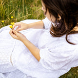 A woman in a white eyelet dress sitting outdoors in a grassy field, holding a small yellow flower.
