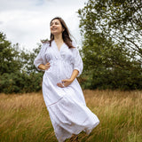A pregnant woman wearing a white long-sleeve maxi dress with a subtle dotted pattern, standing in a field of dry golden grass with trees in the background.