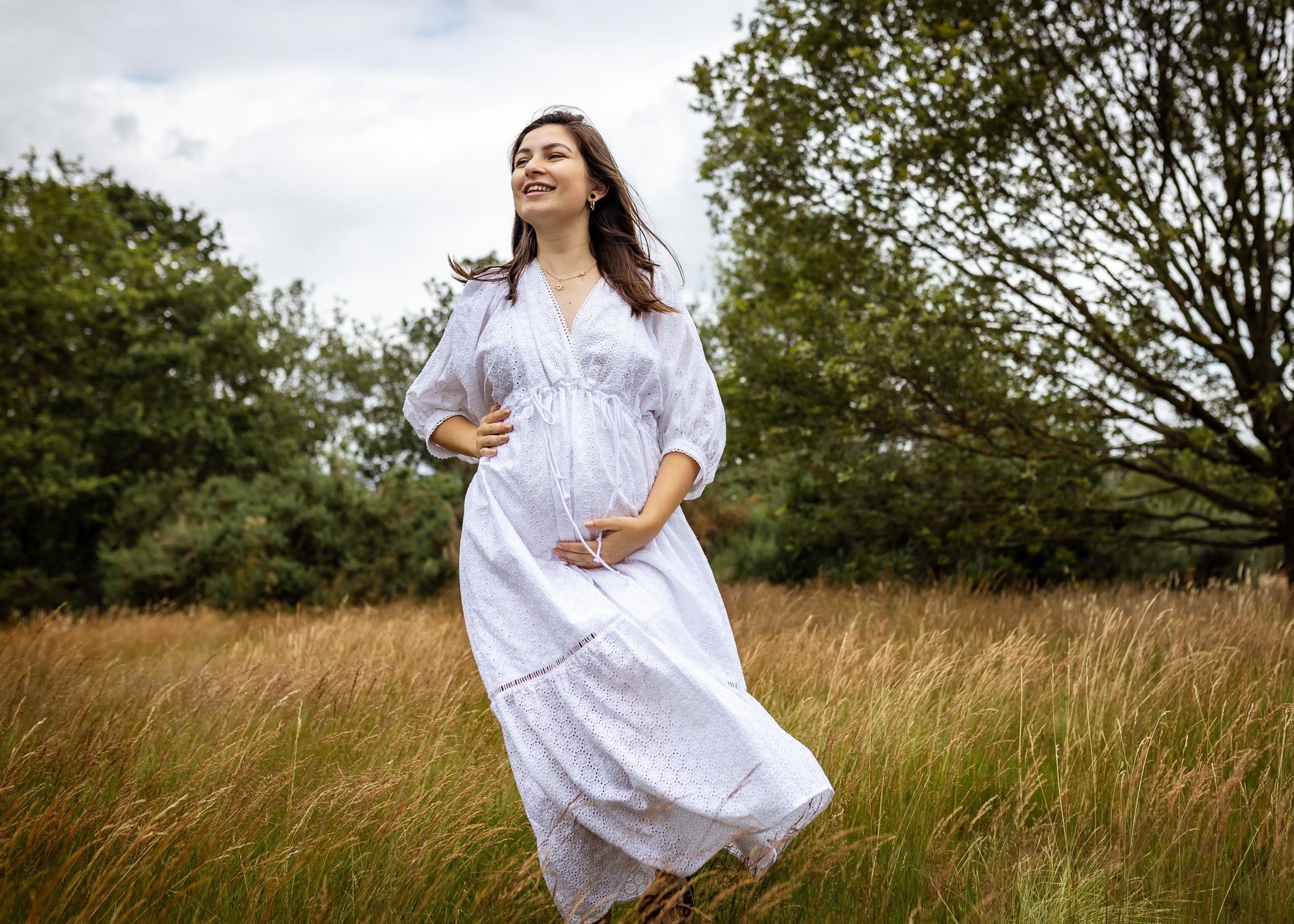 A pregnant woman wearing a white long-sleeve maxi dress with a subtle dotted pattern, standing in a field of dry golden grass with trees in the background.