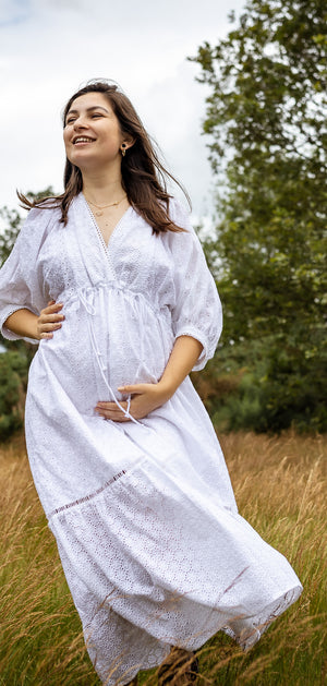 A pregnant woman wearing a white long-sleeve maxi dress with a subtle dotted pattern, standing in a field of dry golden grass with trees in the background.