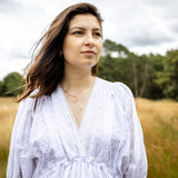 A woman with long brown hair stands in a grassy field wearing a white eyelet long-sleeve blouse. She is looking off-camera with a thoughtful expression, with trees and a cloudy sky in the background.