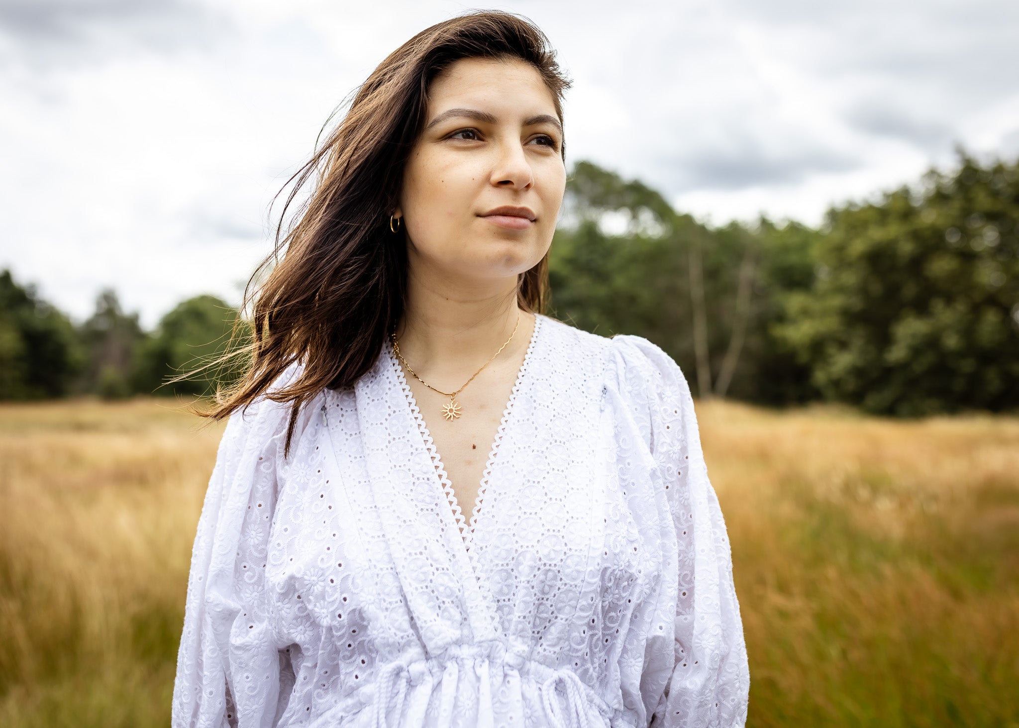A woman with long brown hair stands in a grassy field wearing a white eyelet long-sleeve blouse. She is looking off-camera with a thoughtful expression, with trees and a cloudy sky in the background.