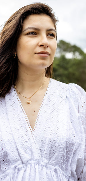 A woman with long brown hair stands in a grassy field wearing a white eyelet long-sleeve blouse. She is looking off-camera with a thoughtful expression, with trees and a cloudy sky in the background.