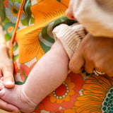 A close-up lifestyle shot of a baby's bare foot resting on a person's lap. The person is wearing a vibrant floral dress with orange, green, and yellow patterns, and holding the baby's leg with one hand while wearing a beige knit sleeve on the baby's other arm.