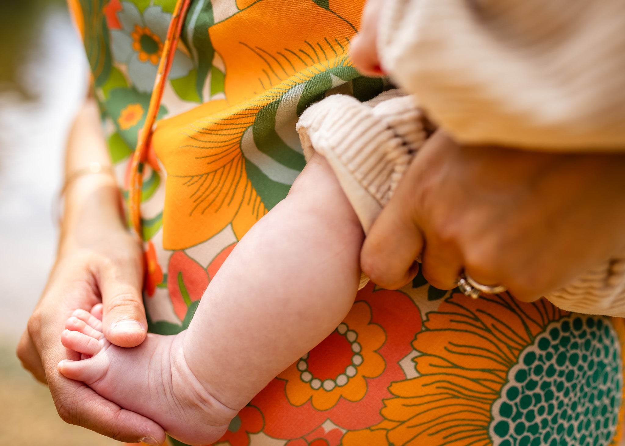 A close-up lifestyle shot of a baby's bare foot resting on a person's lap. The person is wearing a vibrant floral dress with orange, green, and yellow patterns, and holding the baby's leg with one hand while wearing a beige knit sleeve on the baby's other arm.