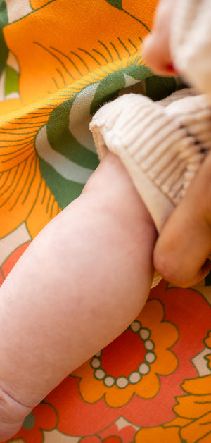 A close-up lifestyle shot of a baby's bare foot resting on a person's lap. The person is wearing a vibrant floral dress with orange, green, and yellow patterns, and holding the baby's leg with one hand while wearing a beige knit sleeve on the baby's other arm.