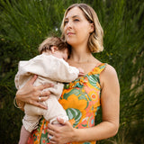 A woman outdoors holding a baby, wearing a sleeveless floral dress with orange, green, and yellow tones. The background consists of blurred green foliage.