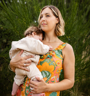 A woman outdoors holding a baby, wearing a sleeveless floral dress with orange, green, and yellow tones. The background consists of blurred green foliage.