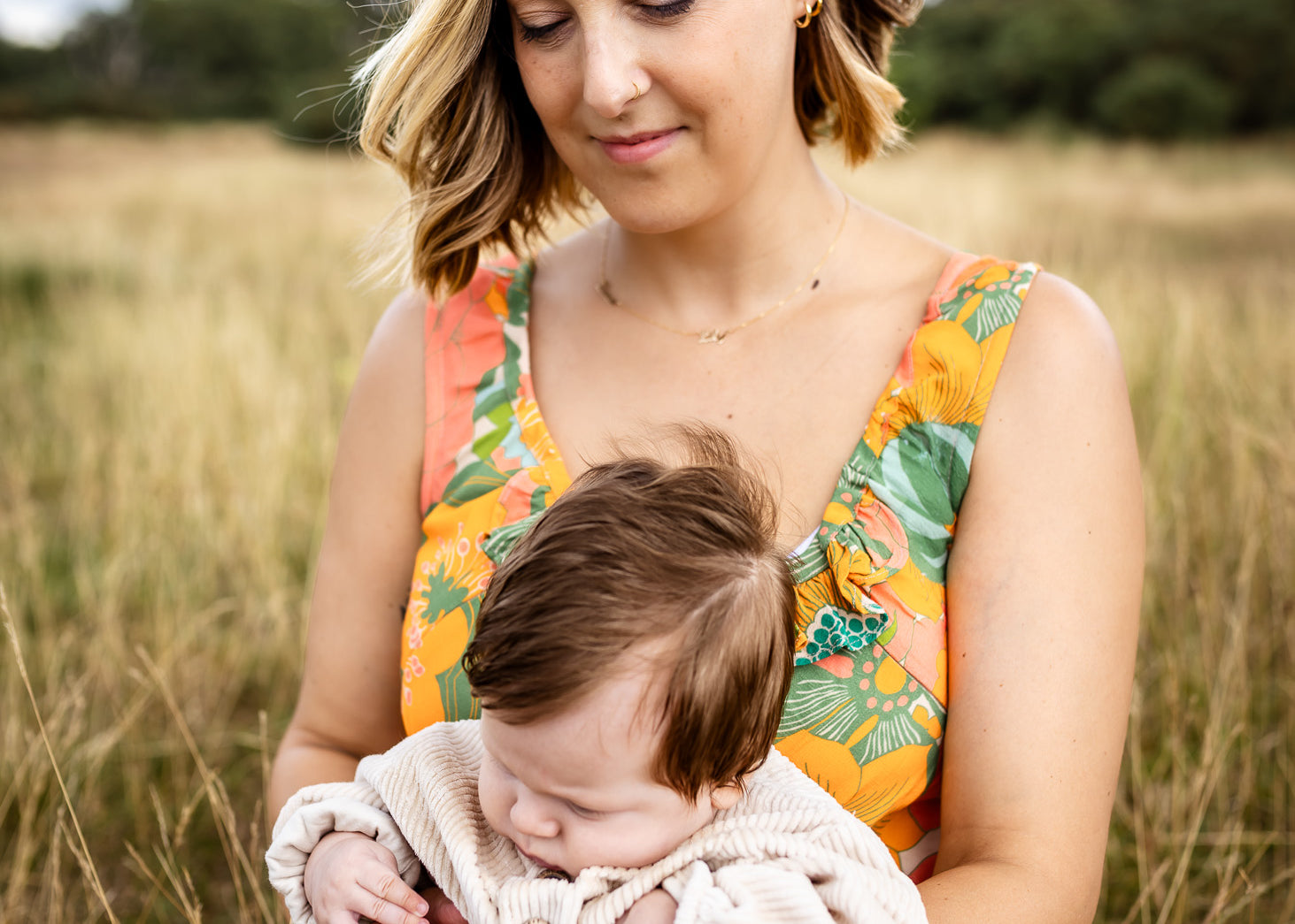 A woman sitting in a grassy field holds a sleeping baby. The woman is wearing a sleeveless floral dress with orange, green, and yellow tones, and the baby is dressed in a light cream knit outfit.
