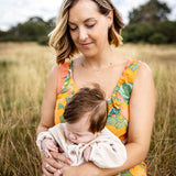 A woman sitting in a grassy field holds a sleeping baby. The woman is wearing a sleeveless floral dress with orange, green, and yellow tones, and the baby is dressed in a light cream knit outfit.