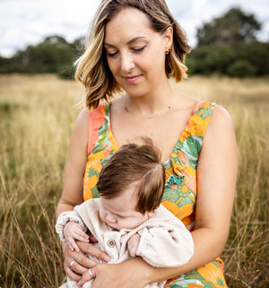 A woman sitting in a grassy field holds a sleeping baby. The woman is wearing a sleeveless floral dress with orange, green, and yellow tones, and the baby is dressed in a light cream knit outfit.