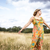 A woman stands in a golden grassy field under a cloudy sky, wearing a sleeveless maxi dress with a vibrant orange, green, and yellow floral pattern.