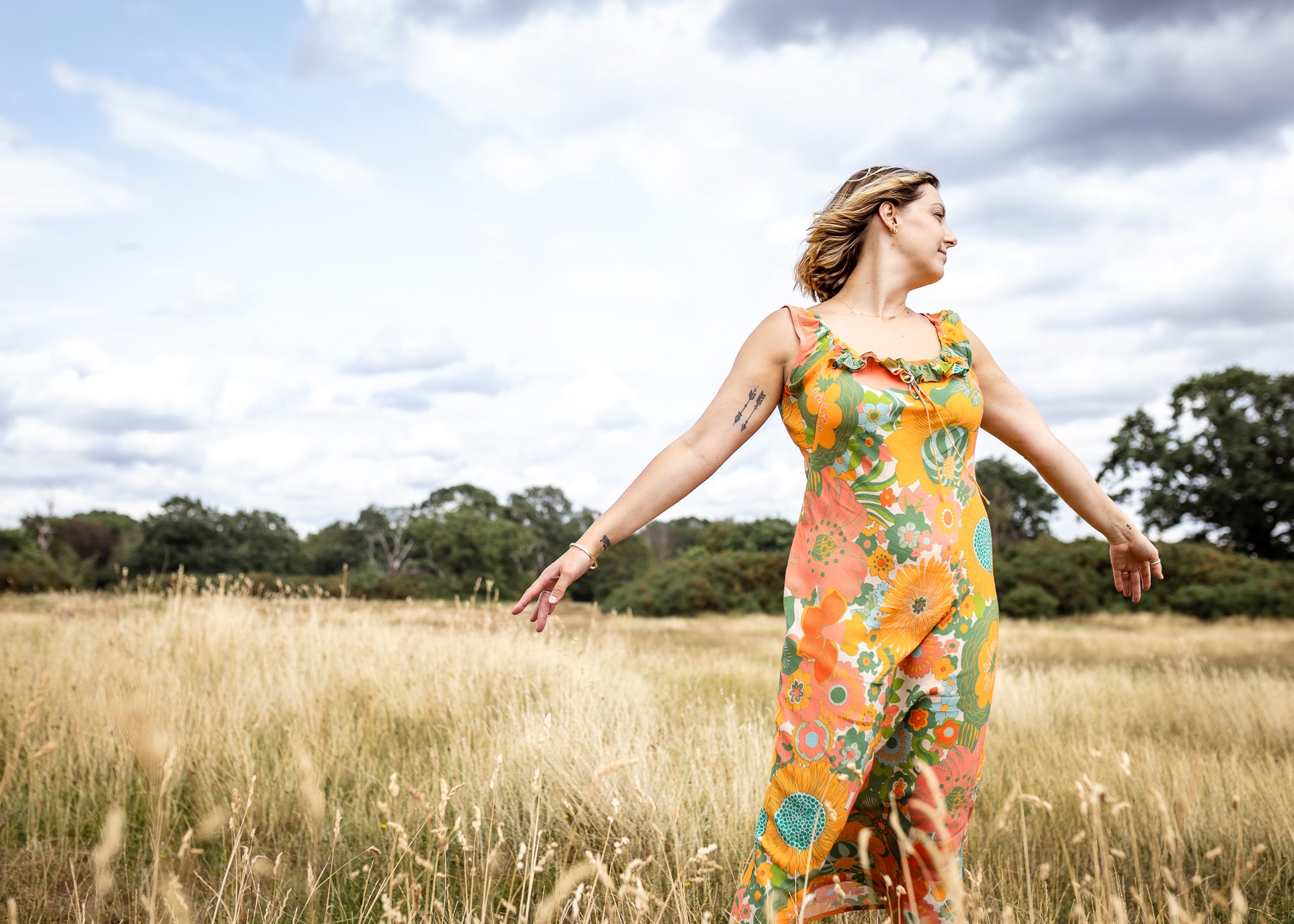 A woman stands in a golden grassy field under a cloudy sky, wearing a sleeveless maxi dress with a vibrant orange, green, and yellow floral pattern.
