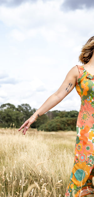 A woman stands in a golden grassy field under a cloudy sky, wearing a sleeveless maxi dress with a vibrant orange, green, and yellow floral pattern.
