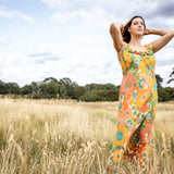 A woman stands in a grassy field wearing a sleeveless maxi dress with a vibrant orange, green, and yellow floral pattern against a backdrop of a cloudy sky and trees.