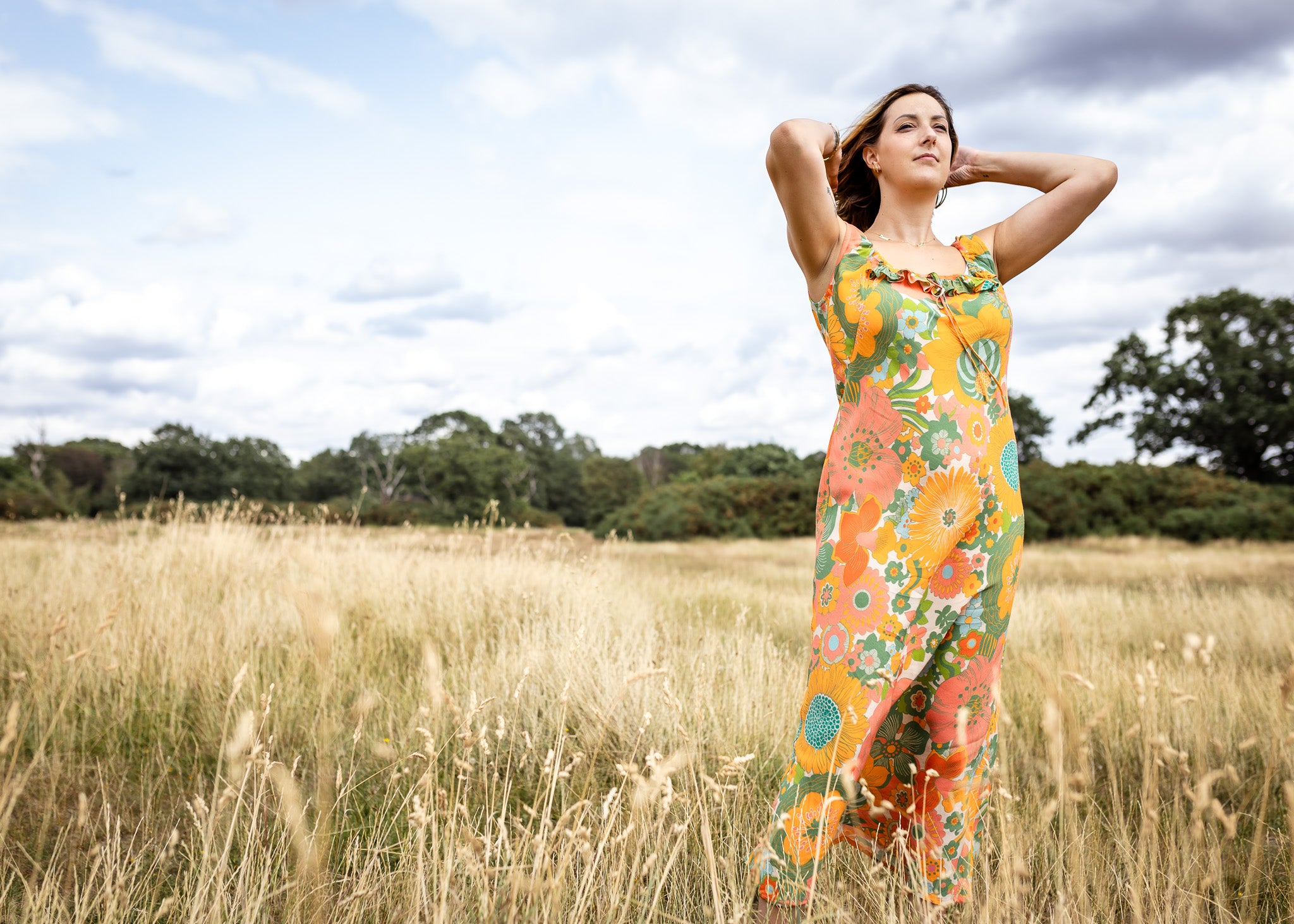A woman stands in a grassy field wearing a sleeveless maxi dress with a vibrant orange, green, and yellow floral pattern against a backdrop of a cloudy sky and trees.
