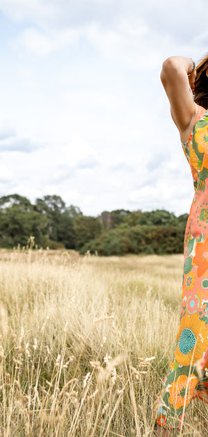 A woman stands in a grassy field wearing a sleeveless maxi dress with a vibrant orange, green, and yellow floral pattern against a backdrop of a cloudy sky and trees.
