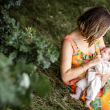 A woman sitting on grass outdoors holds a baby dressed in a light cream outfit. The woman wears a bright orange floral dress, and the scene is framed by green foliage in the foreground.