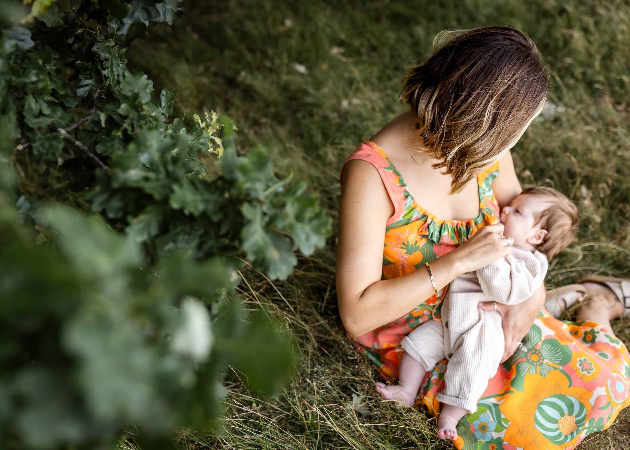 A woman sitting on grass outdoors holds a baby dressed in a light cream outfit. The woman wears a bright orange floral dress, and the scene is framed by green foliage in the foreground.