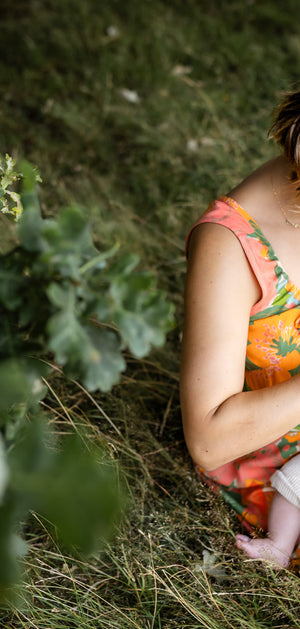 A woman sitting on grass outdoors holds a baby dressed in a light cream outfit. The woman wears a bright orange floral dress, and the scene is framed by green foliage in the foreground.