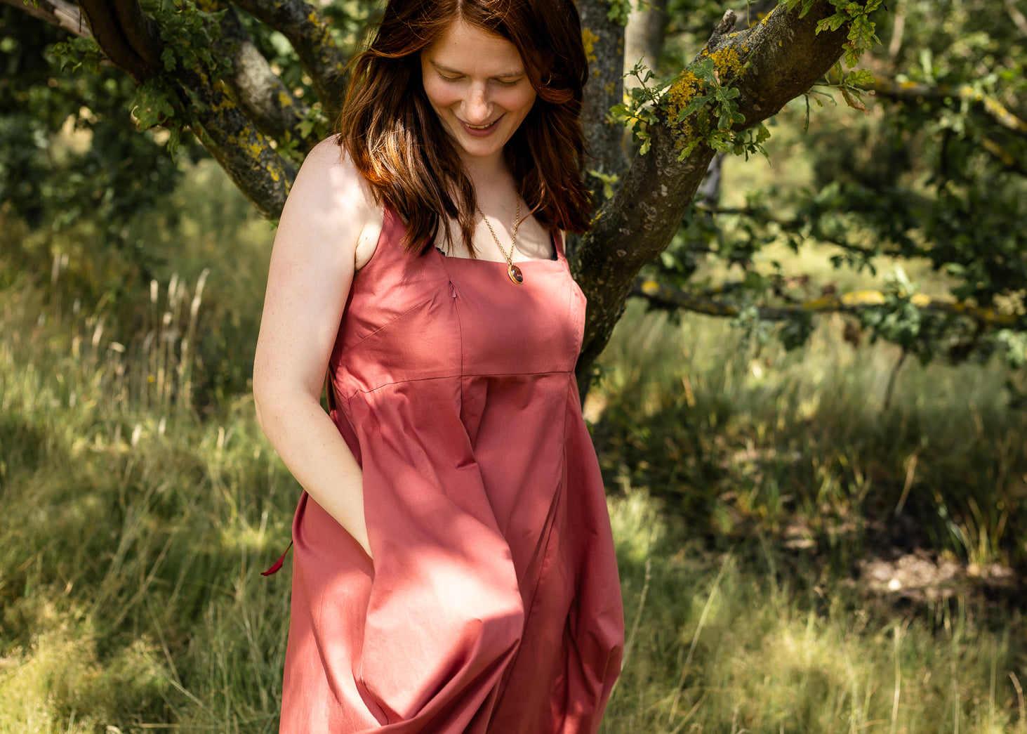 A woman wearing a dusty rose pink sleeveless midi dress stands in a grassy outdoor area under a tree.