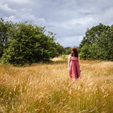 A woman in a pink dress stands in a tall grassy field with trees and a cloudy sky in the background.