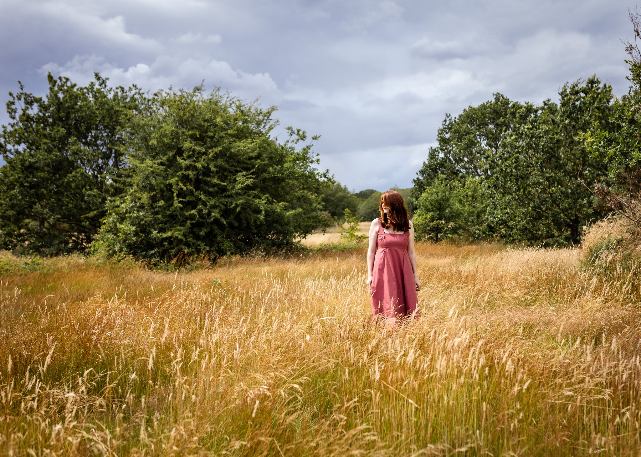 A woman in a pink dress stands in a tall grassy field with trees and a cloudy sky in the background.
