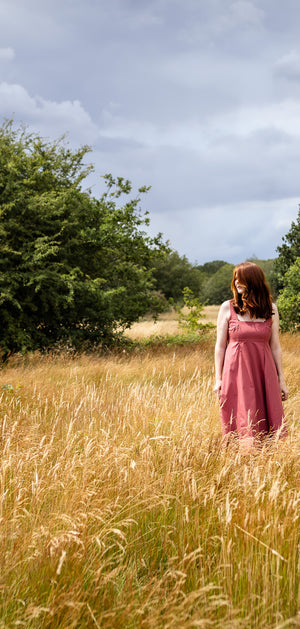 A woman in a pink dress stands in a tall grassy field with trees and a cloudy sky in the background.