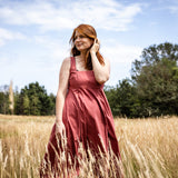 A woman with red hair wearing a dusty rose sleeveless dress stands in a field of dry grass under a blue sky with trees in the background.