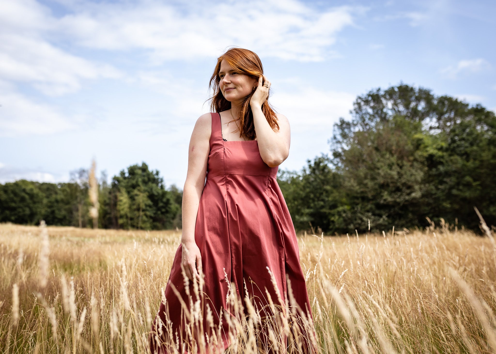 A woman with red hair wearing a dusty rose sleeveless dress stands in a field of dry grass under a blue sky with trees in the background.
